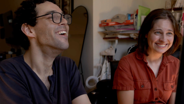 Director Reid Davenport, a white man with curly, short brown hair and glasses, and producer Colleen Cassingham, a white woman with a brown bob wearing an orange jumpsuit, laugh together as they look toward a computer screen, which is just out of the shot. In the background, is an apartment with a full length mirror and an array of books on a shelf.