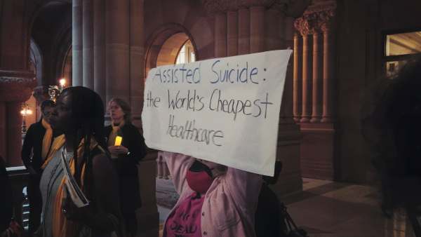 A person holds a sign that reads, ‘ASSISTED SUICIDE: The World’s Cheapest Healthcare.’ They are a wheelchair user with two face masks on, wearing a pink shirt that says ‘NOT DEAD YET,’ in the middle of a busy room at a protest at the New York State Assembly.